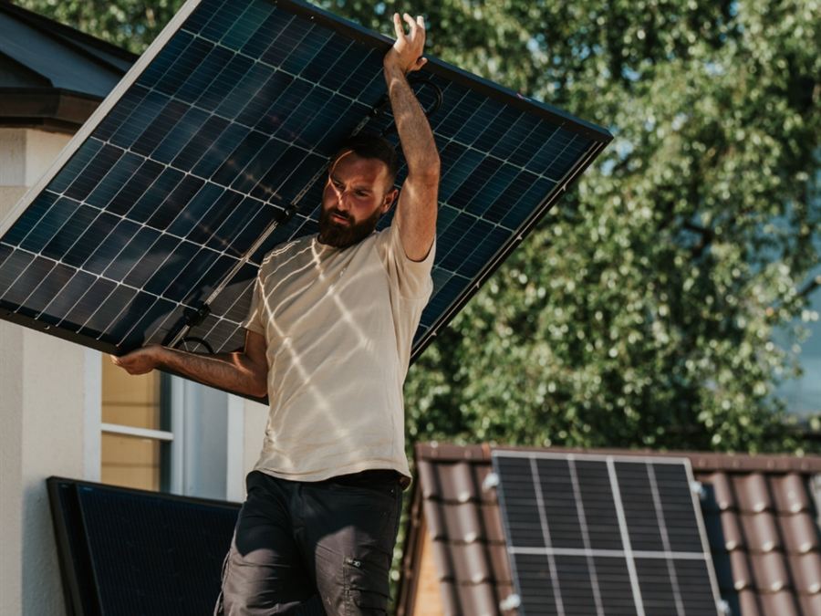 Solaris-Monteur trägt ein Solarmodul über ein Dach, im Hintergrund fertige PV-Anlagen